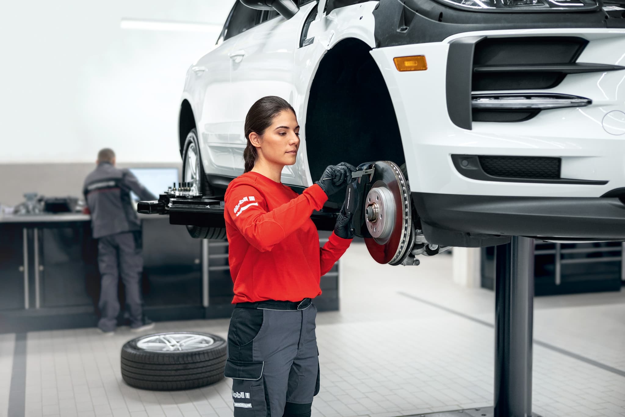 A female mechanic in a red shirt works on a car's brake system in a garage, with another mechanic in the background. A tire is on the floor.